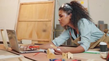 Marangoz Amerikalı siyah kadın kıvırcık saç eskizi yapıyor ve masa başında not alıyor. Ahşap marangozluk atölyesinde dizüstü bilgisayarla, genç bayan öğrenme projesi İnternet 'te marangozhanede.