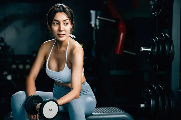 A sporty young woman sitting on a bench at the gym, practicing weightlifting with dumbbells to lift her bicep muscles and improve her physical strength and health, fitness GYM dark background