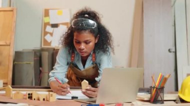 Marangoz Amerikalı siyah kadın kıvırcık saç eskizi yapıyor ve masa başında not alıyor. Ahşap marangozluk atölyesinde dizüstü bilgisayarla, genç bayan öğrenme projesi İnternet 'te marangozhanede.
