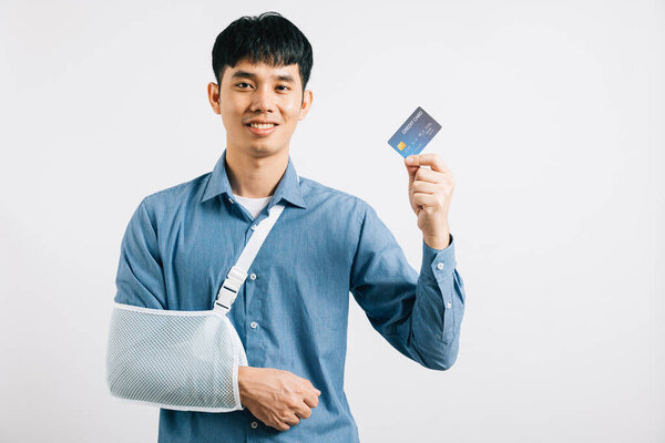 Уверенный в себе человек улыбается через сломанную руку, используя кредитку, чтобы покрыть неотложные медицинские расходы. Счастливый азиат с правой рукой поддержки, изолированные на белом фоне, означающие качество медицинской помощи.