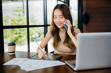 Asian businesswoman sitting at cafe calling mobile phone talking with customer asking about something, smiling female working laptop computer and writing take note on digital tablet at home office