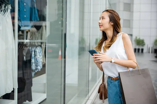 バッグとスマートフォンを持つ幸せな女性は 晴れた日にファッションストアの外に立っています 彼女は技術の利便性とショッピングの喜びを楽しんでいる — ストック写真