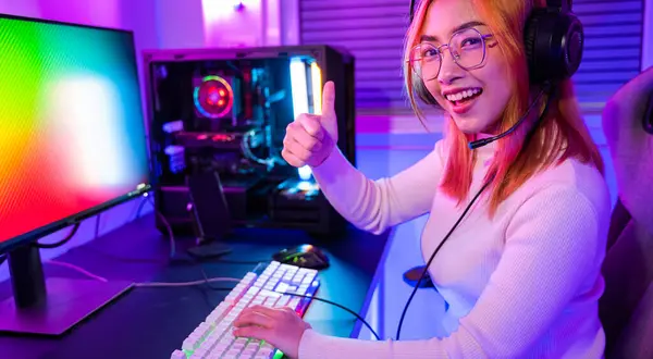 Smiling woman live stream she play video game at home neon lights living room, Gamer playing online game wear gaming headphones looking to camera expressing success with game giving thumbs up sign