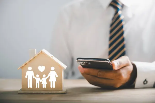 Insuring family well-being and security. Businessman holding mobile phone with family silhouette on wood house. Icons for family, life, health, and home insurance. Reflecting insurance concept.