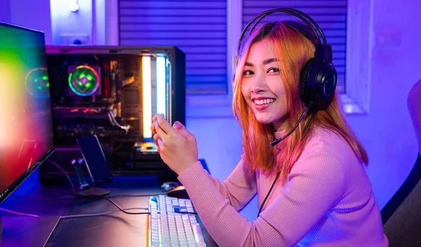 Smiling Asian woman live stream she play video game via smartphone at home neon lights living room, Gamer playing online game application on mobile phone wear gaming headphones looking to camera