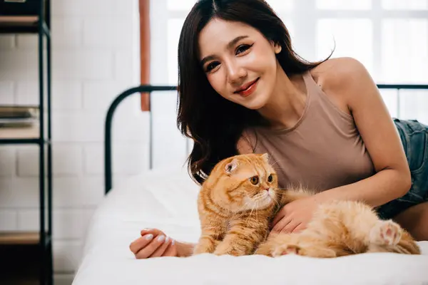 A woman and her orange Scottish Fold cat find happiness and relaxation on the bed in their room. Their bond radiates warmth and care, exemplifying the beauty of pet ownership. Pat love