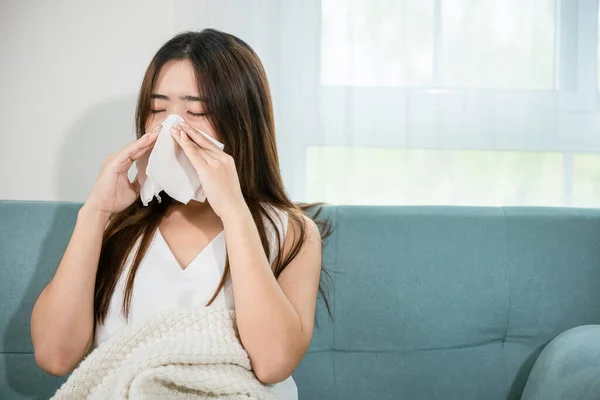 Sick woman sitting under blanket on sofa and sneeze with tissue paper in living room, Asian young female blowing nose sneezing in tissue at home, fever caught cold