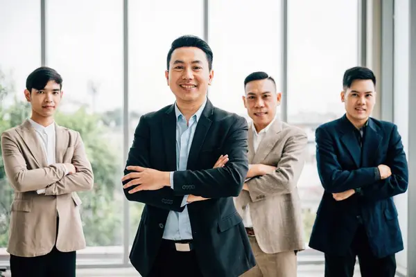 In a creative office, a smart casual-clad businessman smiles with confidence, arms crossed. Diverse Asian male and female colleagues stand together at a startup, underlining teamwork and success.