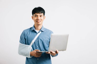 Kırık bir kola rağmen, Asyalı bir işadamı atel takıyor, dizüstü bilgisayar tutuyor ve özveriyle çalışıyor. Stüdyo çekimi beyaz arkaplanda izole edildi, dayanıklılık ve iyileşme göstergesi.