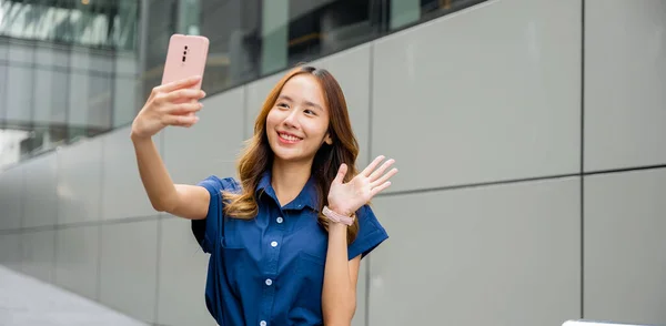 Smiling young lady taking a selfie with her cell phone in the city, showcasing her trendy style and accessories. Happy woman using mobile technology for personal branding and social media.