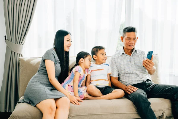 A joyful family shares laughter watching videos on a smartphone while seated on a couch. Portraying familial happiness bonding and shared technology enjoyment among parents and children.