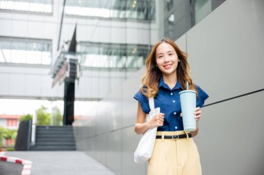 Asyalı güzel bir iş kadını elinde çelik termos bardak bardak tutarak gülümsüyor modern bina ofisinin yakınındaki sokakta yürüyor mutlu bir kadın kameraya bakıyor.