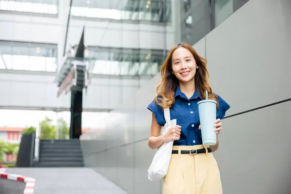 Asian beautiful business woman confident smiling with cloth bag holding steel thermos tumbler mug water glass she walking outdoors on street near modern building office, Happy female looking to camera