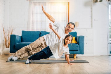 Bir baba ve kızı, evde yoga yaparken uyumlu bir aile anı yaratırlar. Mutluluk ve kas gücünü vurgularlar. Gülümsemeleri pozitiflik yayar..