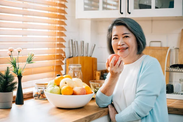 In the kitchen, a happy old woman, a grandmother, enjoys cooking and preparing healthy vegan food. She selects a fresh apple to eat, embodying the diet concept. A portrait of happiness and wellness.