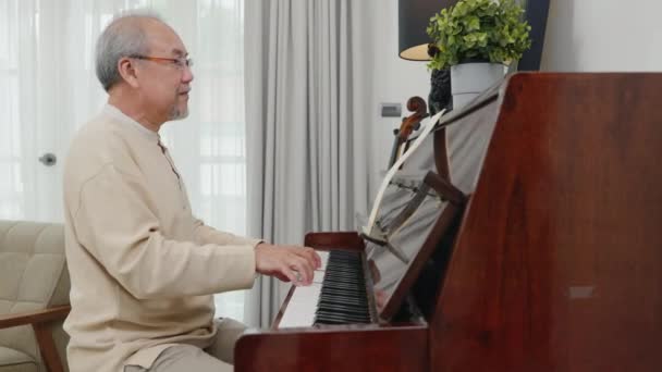 幸せなリラクゼーション眼鏡をかけたアジアの高齢者は自宅でピアノを弾くことを学びます 高齢者はリビングルームでピアノで音楽を演奏します 退職後のライフスタイル 手を閉じる — ストック動画