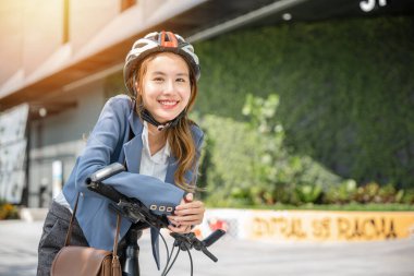 Şehirde Asyalı bir iş kadını, kask takmış ve takım elbiseli bisikletli neşeli bir sabah işe gitmek için hazır bekliyor. Bu resim iş ve açık hava eğlencesini birleştirir.