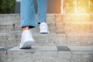Spor ayakkabılarıyla, bir kadın şehir merdivenine çıkıyor, kararlılığını ve gelişimini gösteriyor. Her adım onun başarıya ve kişisel gelişime olan sarsılmaz yolculuğunun bir yansıması. Adım at.