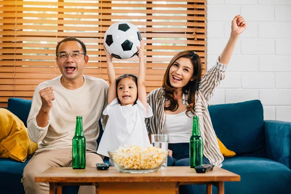 In the comfort of their home, a happy family cheers for a football match with popcorn and a ball. Their togetherness, shouting, and bonding celebrate the triumph and excitement of the game.
