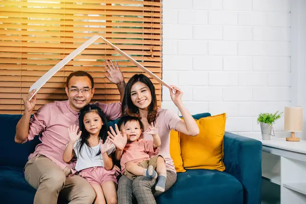 Their new house happy family sits on sofa holding cardboard roof above them symbolizing protection and investment. Laughter togetherness and planning for their relocation at home is what it all about.