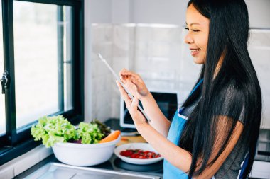 Asyalı bir kadın yemek pişirirken mutlu bir şekilde mutfağında duruyor. Teknoloji aşçılığının ve ev memnuniyetinin kusursuz bütünleşmesini tasvir etmek.