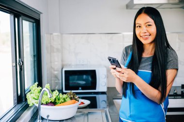 Gülümseyen kadın yemek pişirirken mutfak masasına eğilip cep telefonundaki mesajları okuyor. Memlekette neşeli ev hanımları modern çoklu görev rutinini gözler önüne sermek. cep telefonu