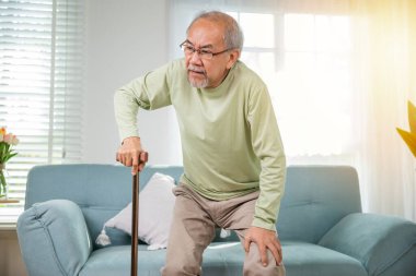 Asyalı, gözlüklü, bastonla kanepeden kalkmak için yazı yazan yaşlı bir adam, baston tutan diz ağrısı çeken yaşlı bir adam, elinde baston tutan yaşlı engelli bir adam.