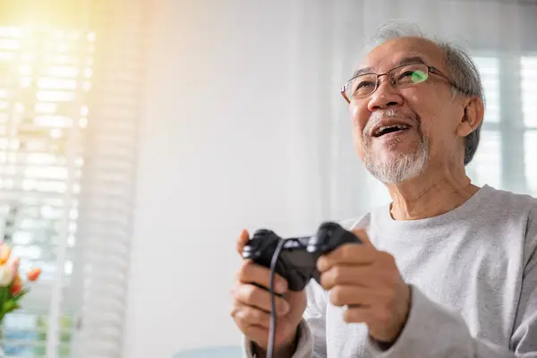 亚洲的成熟男人用游戏控制器玩电子游戏 退休老人笑着坐在沙发上玩人生游戏 老年人喜欢在家里拿着操纵杆玩电子游戏 — 图库照片