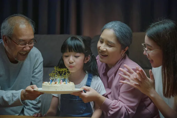 Happy Asian family grandparents and mother celebrating enjoy birthday party with cake of granddaughter Blowing candle at home in night time, Birthday surprise concept
