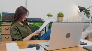 Asyalı genç iş kadını rahat rahat oturup ayakları bilgisayar masasına yaslanıyor rapor belgelerini panoda okuyor, elinde mektupları tutan gülümseyen bir kadın.