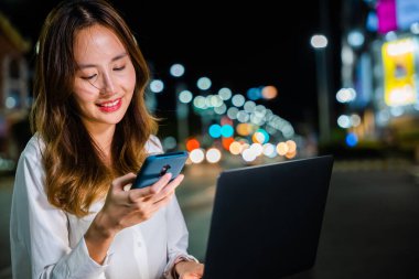 Genç Asyalı kadın gece şehir caddesinde dizüstü bilgisayar kullanırken telefonla konuşuyor, teknolojiyle birden fazla iş yapıyor..