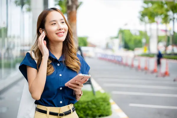 Happy young woman multitasking with wireless earbuds and smartphone in an urban environment. Shes a portrait of a modern entrepreneur.