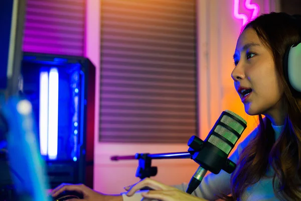 Happy Asian streamer female talking with microphone in gaming room, young gamer woman wear headphones playing video games online on computer she live stream and chat with fans, game streaming concept