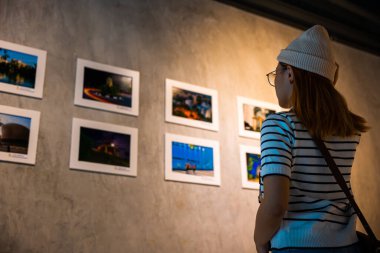 Asyalı kadın renkli çerçeveli resimlerin önünde dikilip beyaz duvara bakıyor, insanlar fotoğraf çerçevesinde sergi sanat galerisine yaslanıyor,