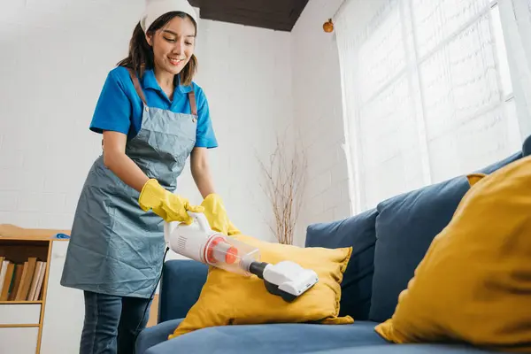 陽気な性質を持つアジアのハウスキーパーは リビングルームのソファをきれいにする掃除機を使用しています 彼女の衛生と家具のケアに焦点を当てることは 彼女の専用の家事の一部です — ストック写真