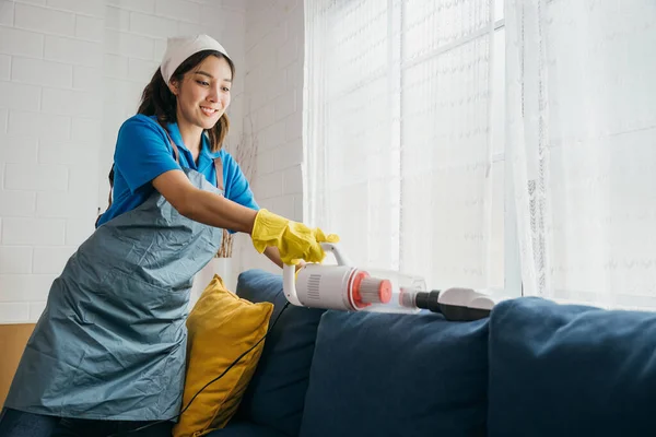 快適なリビングルームでは アジアの女性メイドがソファをきれいにする掃除機を使用しています 衛生と家具ケアへの彼女の献身は 家事への近代的なアプローチを反映しています — ストック写真