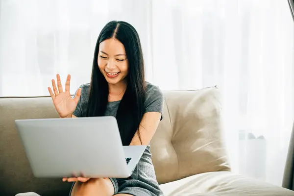 Young woman at laptop waves during video call smiling. Teacher makes hello gesture welcoming clients. Engaged in distant meeting fostering connections and communication.