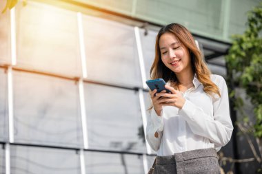 Asyalı iş kadını elinde cep telefonuyla ofis dışında mesaj atıyor, şehir caddesinde akıllı telefon kullanan mutlu genç kadın, cep telefonu uygulamaları, sosyal medyayı kontrol ediyor.