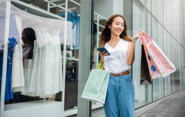 Alışveriş poşeti ve akıllı telefonu elinde tutan mutlu Asyalı güzel bir kadın alışveriş merkezinin penceresinin yanında gülümseyen müşteri kız yaşam tarzı şehrin caddesinde alışveriş yaptıktan sonra cep telefonunu kullanıyor.