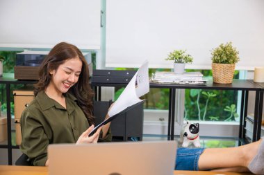 Asyalı genç iş kadını rahat rahat oturup ayakları bilgisayar masasına yaslanıyor rapor belgelerini panoda okuyor, elinde mektupları tutan gülümseyen bir kadın.