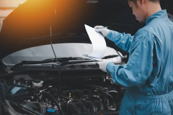 青いワークウェアのアジアのメカニックは 自動車修理工場で車を修理しながら 紙に書いています クローズアップショット — ストック写真