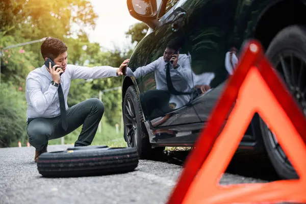 Broken down car with a red triangle warning sign on the road. Businessman waiting for assistance while leaning on the car wheel. Concept of car breakdown and roadside rescue service.