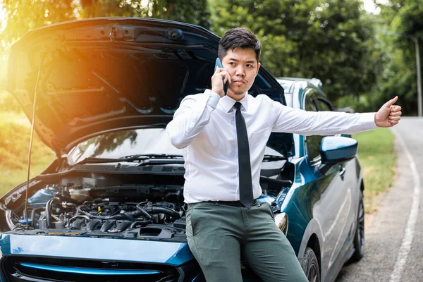 Asian businessman stranded on the road after car breakdown, using phone to call for help. Candid shot of man waiting for assistance and giving thumbs up. Transportation and insurance concept.