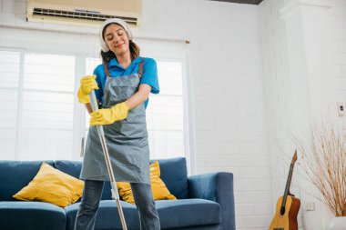 Asyalı neşeli hizmetçi, kulaklıkla şarkı söylüyor. Neşeli meşguliyet müzik heyecanıyla kaynaştı. Teknolojiyle modern ev işi titreşimi. Bana bir şarkı söyle, Happy and Fun during Cleaning