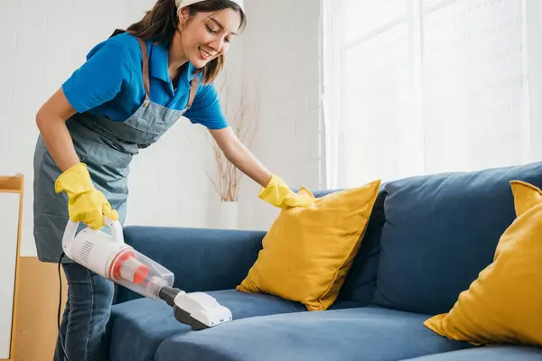 アジアのメイドが陽気な掃除機でリビングルームのソファーを掃除しています 家具ケアのための衛生と現代のクリーニング技術への彼女の献身は明らかです — ストック写真