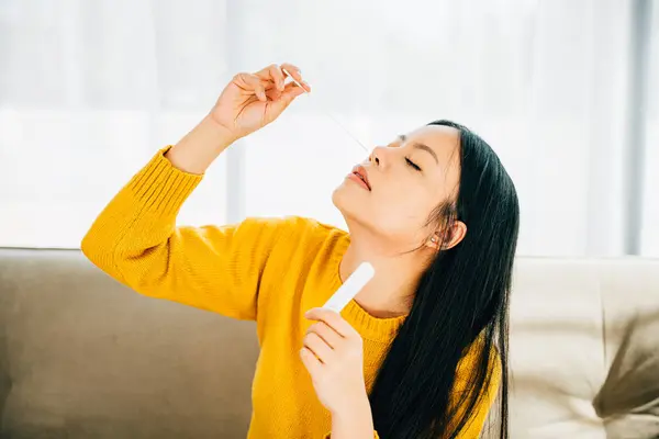 An adult Asian woman self-tests for COVID-19 at home inserting a swab into her nose. Reflecting virus prevention testing and measures during the coronavirus pandemic.