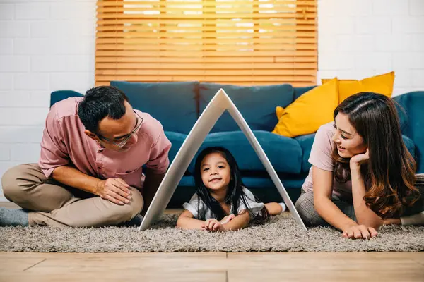 A happy family on a couch holding a cardboard roof in their new home symbolizing safety and support. Asian parents and their daughter embody affectionate togetherness.