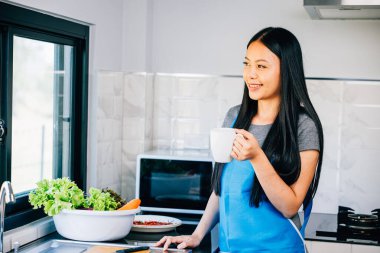 Kaygısız bir kadın sabah kahvesinin tadını çıkarırken rahat bir mutfakta gülümsüyor. Neşeli, rahat bir anı resmetmek modern bir evde sıcak bir içkinin tadını çıkarmak..