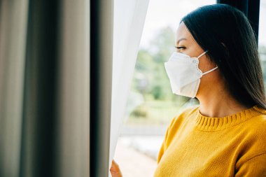 COVID-19 sıkıntısını ifade eden maskeli kadın üzgün ve yalnız hissederek pencereden dışarı bakıyor. Korku yalnızlığını ve salgın krizinin etkisini yansıtıyor..