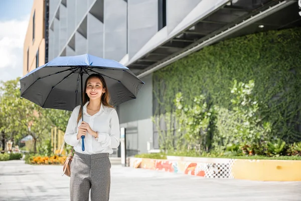 スコッチングの日にオフィスに歩いていると 若いビジネスマンは暑い日差しから身を守るために傘を持っています 彼女の決意と汗は 成功への彼女のコミットメントを強調しています — ストック写真